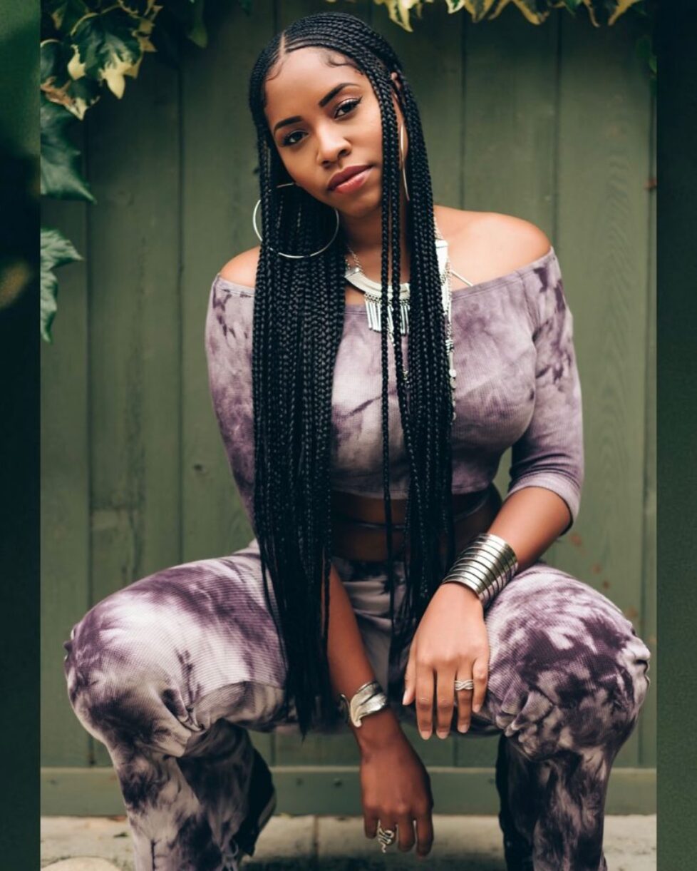 A woman in a purple tie-dye outfit poses with long braided hair against a green background.